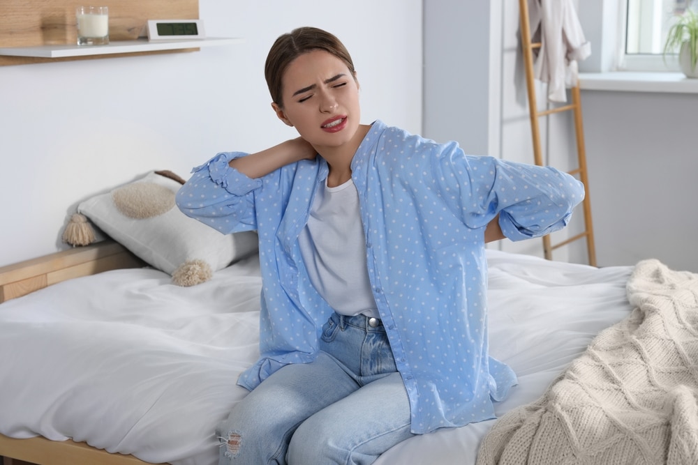 Woman sitting on bed holding her neck and back, dealing with chronic pain that could be addressed with red light therapy