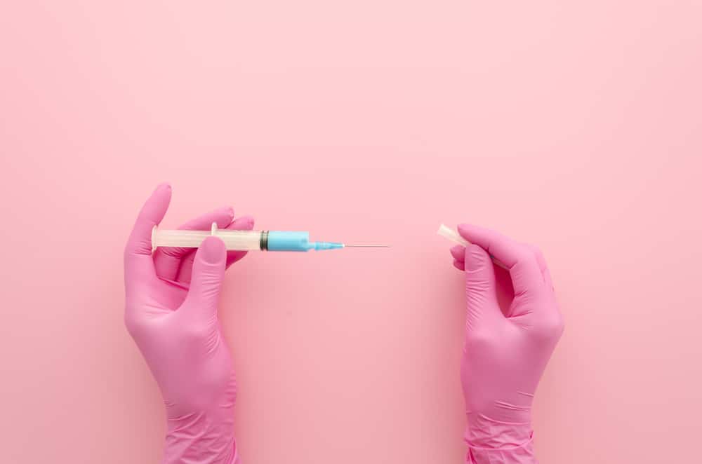 Gloved hand holding a blue vitamin shot syringe over a pink background.